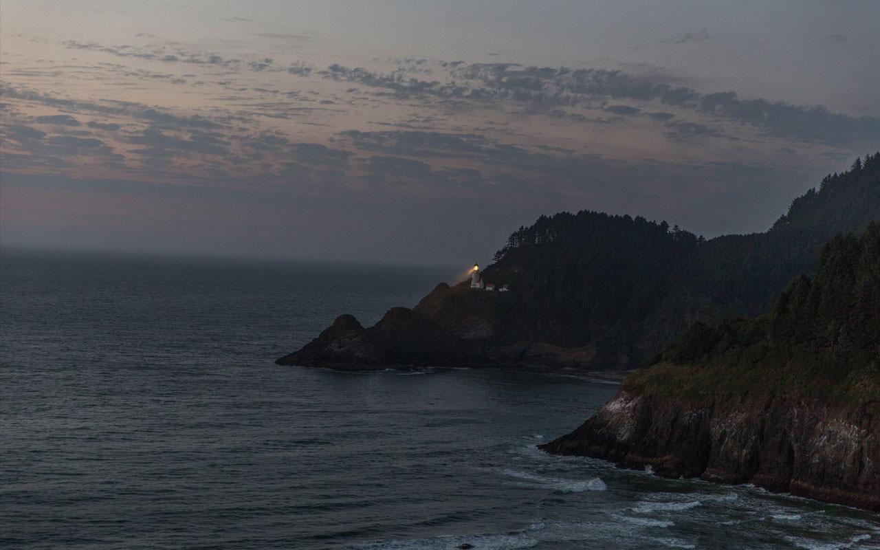 Photo caption: Foreground, waves crash along the shoreline near an Oregon coast cliff. Background, rays of light radiate from a coastal lighthouse into the Pacific Ocean. Photo credit: Jamie Francis Photography.