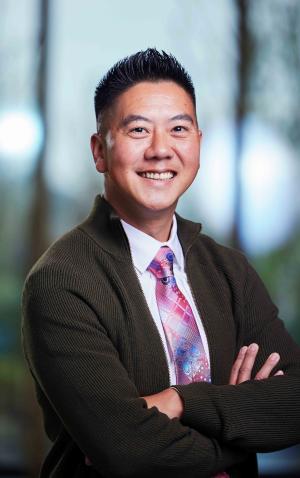 A portrait of Huy in a dark green sweater and colorful tie standing in front of a window