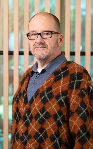 A photo of Aaron wearing an orange argyle sweater front of a wall of wood slats
