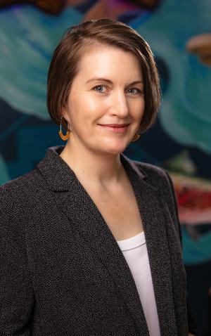 A portrait of Erin in a grey blazer standing in front of a vibrant mural