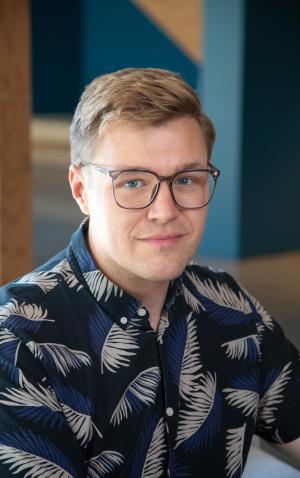 A portrait of Tyler in a blue shirt standing in front of a wooden wall